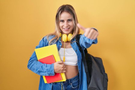 Téléchargez les photos : Jeune femme blonde portant un sac à dos étudiant et tenant des livres approuvant faire un geste positif avec la main, les pouces levés souriant et heureux pour le succès. geste gagnant. - en image libre de droit