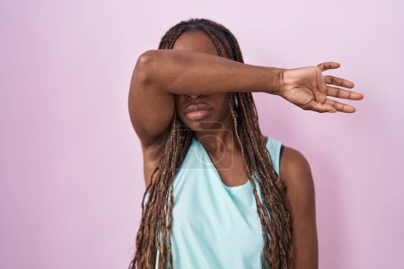 Téléchargez les photos : Femme afro-américaine debout sur fond rose couvrant les yeux avec le bras, l'air grave et triste. concept de non-visibilité, de dissimulation et de rejet - en image libre de droit
