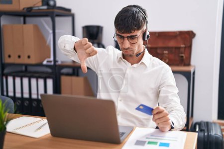 Photo for Young hispanic man working using computer laptop holding credit card looking unhappy and angry showing rejection and negative with thumbs down gesture. bad expression. - Royalty Free Image