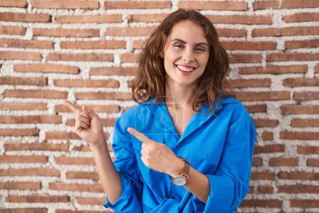 Téléchargez les photos : Belle femme brune debout sur un mur de briques souriant et regardant la caméra pointant avec deux mains et doigts sur le côté. - en image libre de droit