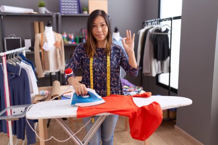 Téléchargez les photos : Moyen âge femme chinoise couturière designer repassage vêtements faire ok signe avec les doigts, sourire geste amical excellent symbole - en image libre de droit