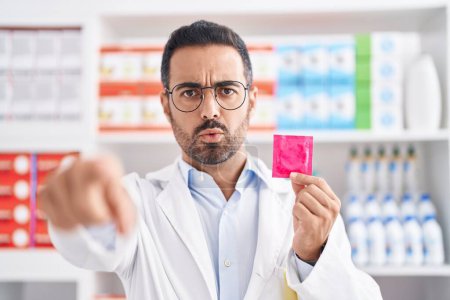 Téléchargez les photos : Homme hispanique avec barbe travaillant à la pharmacie pharmacie tenant préservatif pointant du doigt la caméra et à vous, geste confiant regardant sérieux - en image libre de droit