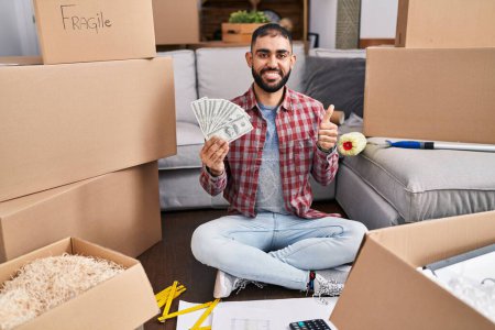 Photo for Middle east man with beard sitting on the floor at new home holding money smiling happy and positive, thumb up doing excellent and approval sign - Royalty Free Image