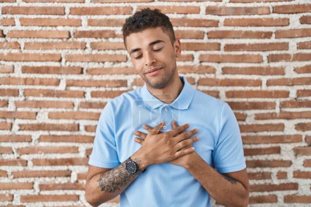 Photo for Brazilian young man standing over brick wall smiling with hands on chest with closed eyes and grateful gesture on face. health concept. - Royalty Free Image