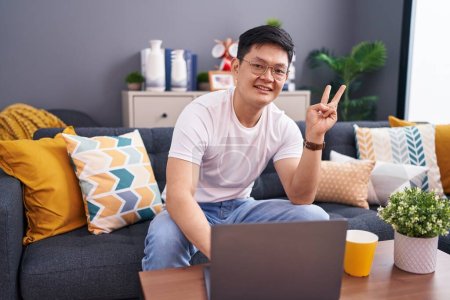 Photo for Young asian man using laptop at home sitting on the sofa smiling with happy face winking at the camera doing victory sign with fingers. number two. - Royalty Free Image