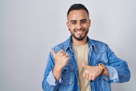 Téléchargez les photos : Jeune homme hispanique debout sur fond isolé pointant le dos derrière avec la main et les pouces levés, souriant confiant - en image libre de droit
