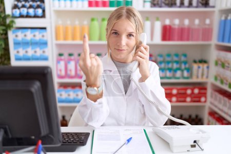 Photo for Young caucasian woman working at pharmacy drugstore speaking on the telephone showing middle finger, impolite and rude fuck off expression - Royalty Free Image