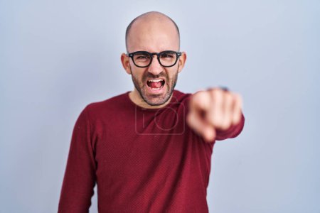 Téléchargez les photos : Jeune homme chauve avec la barbe debout sur fond blanc portant des lunettes pointant mécontent et frustré à la caméra, en colère et furieux contre vous - en image libre de droit