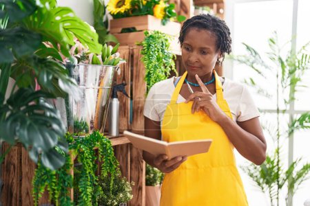 Photo for Middle age african american woman florist reading notebook with doubt expression at flower shop - Royalty Free Image