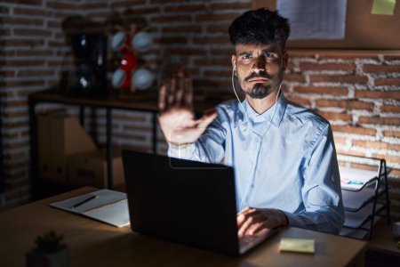 Photo for Young hispanic man with beard working at the office at night doing stop sing with palm of the hand. warning expression with negative and serious gesture on the face. - Royalty Free Image