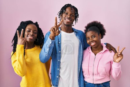 Téléchargez les photos : Groupe de trois jeunes Noirs debout ensemble sur fond rose souriant avec un sourire heureux clin d'oeil à la caméra faisant signe de victoire avec les doigts. numéro deux. - en image libre de droit