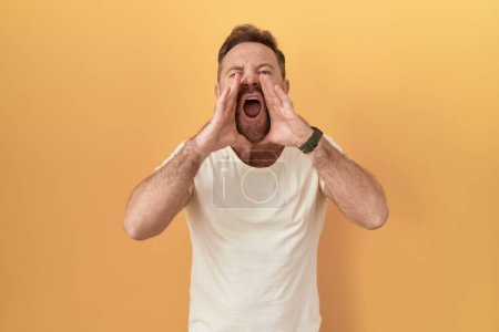 Middle age man with beard standing over yellow background shouting angry out loud with hands over mouth 