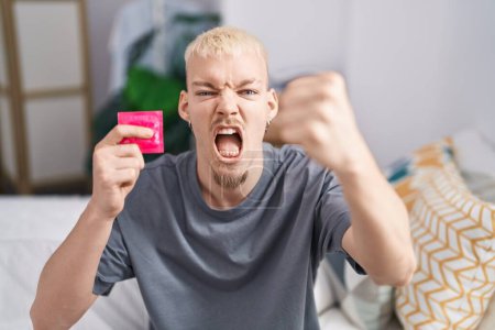 Téléchargez les photos : Jeune homme caucasien tenant préservatif assis sur le lit ennuyé et frustré criant de colère, criant fou de colère et la main levée - en image libre de droit