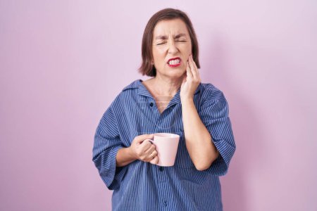 Foto de Mujer hispana de mediana edad bebiendo una taza de café tocando la boca con la mano con expresión dolorosa debido a dolor de muelas o enfermedad dental en los dientes. concepto de dentista. - Imagen libre de derechos