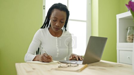 Foto de Mujer afroamericana usando la escritura del ordenador portátil en el cuaderno en casa - Imagen libre de derechos