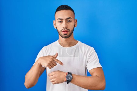 Téléchargez les photos : Young hispanic man standing over blue background in hurry pointing to watch time, impatience, upset and angry for deadline delay - en image libre de droit