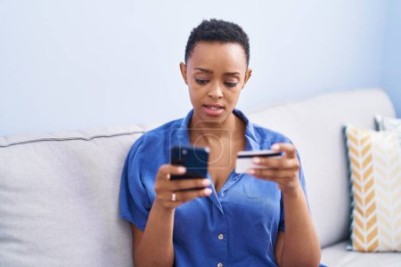 Foto de Mujer afroamericana usando teléfono inteligente y tarjeta de crédito sentada en el sofá en casa - Imagen libre de derechos
