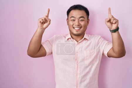 Photo for Chinese young man standing over pink background smiling amazed and surprised and pointing up with fingers and raised arms. - Royalty Free Image