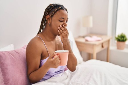 Téléchargez les photos : Femme afro-américaine assise sur le lit bâillant et buvant du café dans la chambre - en image libre de droit
