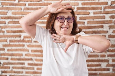 Téléchargez les photos : Femme âgée avec des lunettes debout sur le mur de briques souriant joyeux jeu jeter un coup d'oeil un boo avec les mains montrant le visage. surpris et sortis - en image libre de droit