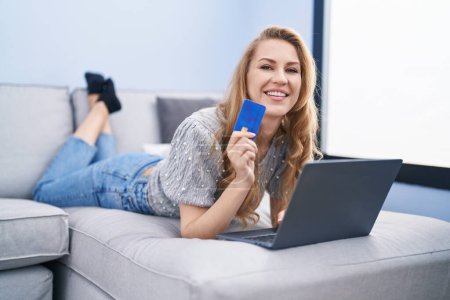 Téléchargez les photos : Jeune femme blonde utilisant un ordinateur portable et carte de crédit couché sur le canapé à la maison - en image libre de droit
