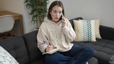 Téléchargez les photos : Jeune femme blonde écrit sur ordinateur portable parler sur smartphone à la maison - en image libre de droit