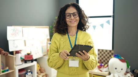 Téléchargez les photos : Moyen âge hispanique femme maternelle professeur souriant confiant en utilisant touchpad à la maternelle - en image libre de droit