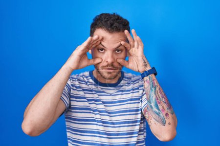 Photo for Young hispanic man standing over blue background trying to open eyes with fingers, sleepy and tired for morning fatigue - Royalty Free Image