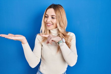 Téléchargez les photos : Jeune femme caucasienne debout sur fond bleu étonné et souriant à la caméra tout en présentant avec la main et pointant du doigt. - en image libre de droit