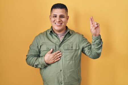 Téléchargez les photos : Jeune homme hispanique debout sur fond jaune souriant jurer avec la main sur la poitrine et les doigts vers le haut, faisant un serment de fidélité promesse - en image libre de droit