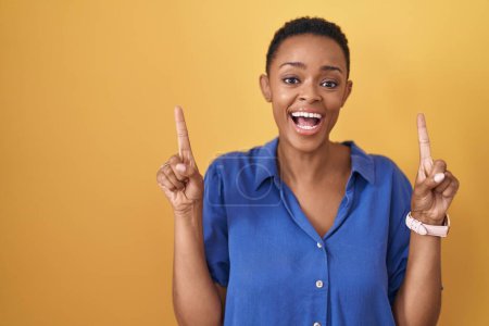 Téléchargez les photos : Femme afro-américaine debout sur fond jaune souriant étonné et surpris et pointant vers le haut avec les doigts et les bras levés. - en image libre de droit