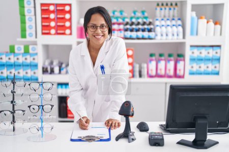 Photo for Young hispanic woman pharmacist writing on document using computer at pharmacy - Royalty Free Image