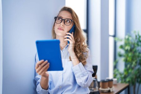 Téléchargez les photos : Jeune femme blonde travailleuse d'affaires parlant sur le smartphone en utilisant le pavé tactile au bureau - en image libre de droit