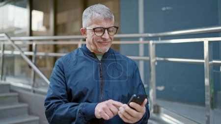 Foto de Hombre de pelo gris de mediana edad sonriendo confiado usando el teléfono inteligente en la calle - Imagen libre de derechos