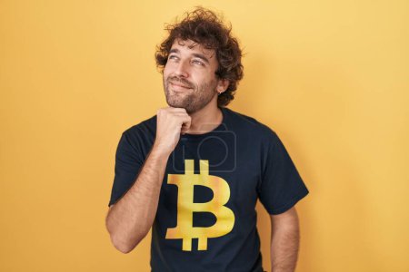 Photo for Hispanic young man wearing bitcoin t shirt with hand on chin thinking about question, pensive expression. smiling with thoughtful face. doubt concept. - Royalty Free Image