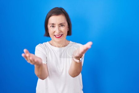 Foto de Middle age hispanic woman standing over blue background smiling cheerful offering hands giving assistance and acceptance. - Imagen libre de derechos