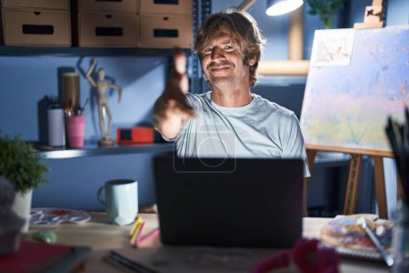 Foto de Hombre de mediana edad sentado en el estudio de arte con el ordenador portátil por la noche sonriendo amable ofreciendo apretón de manos como saludo y bienvenida. negocios exitosos. - Imagen libre de derechos