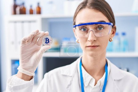 Photo for Young caucasian woman working at scientist laboratory holding bitcoin thinking attitude and sober expression looking self confident - Royalty Free Image