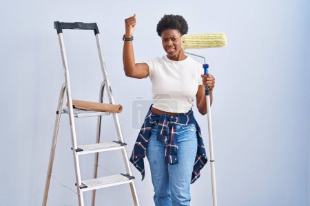 Photo for African american woman holding roller painter angry and mad raising fist frustrated and furious while shouting with anger. rage and aggressive concept. - Royalty Free Image