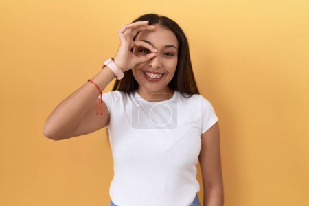 Photo for Young arab woman wearing casual white t shirt over yellow background doing ok gesture with hand smiling, eye looking through fingers with happy face. - Royalty Free Image