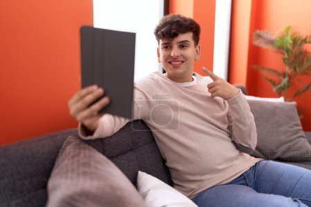 Foto de Joven hombre no binario usando touchpad sentado en el sofá sonriendo feliz señalando con la mano y el dedo - Imagen libre de derechos