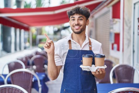 Photo for Arab man with beard wearing waiter apron at restaurant terrace smiling happy pointing with hand and finger to the side - Royalty Free Image