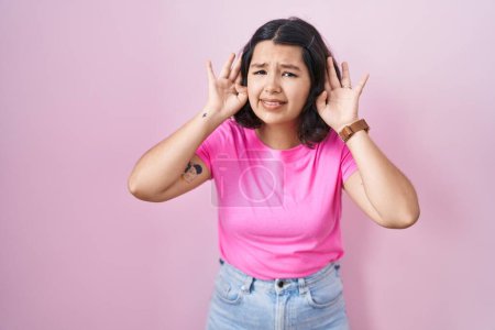 Téléchargez les photos : Jeune femme hispanique debout sur fond rose essayant d'entendre les deux mains sur le geste de l'oreille, curieuse de commérages. problème auditif, sourd - en image libre de droit
