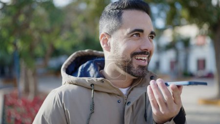 Foto de Joven hombre hispano sonriendo confiado enviando mensaje de voz por smartphone en park - Imagen libre de derechos