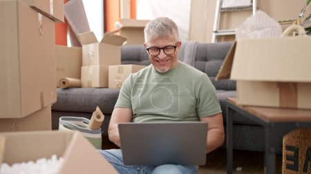 Téléchargez les photos : Moyen âge homme aux cheveux gris utilisant ordinateur portable assis sur le sol à la nouvelle maison - en image libre de droit