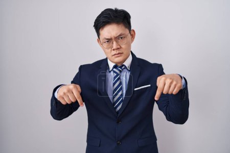 Téléchargez les photos : Jeune homme asiatique portant costume d'affaires et cravate pointant vers le bas regardant triste et contrarié, indiquant la direction avec les doigts, malheureux et déprimé. - en image libre de droit