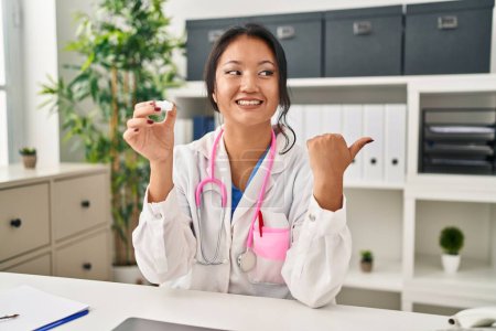 Foto de Joven asiática mujer vistiendo doctor uniforme y estetoscopio apuntando pulgar hasta el lado sonriendo feliz con boca abierta - Imagen libre de derechos
