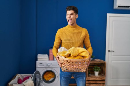 Photo for Young hispanic man holding laundry basket angry and mad screaming frustrated and furious, shouting with anger. rage and aggressive concept. - Royalty Free Image