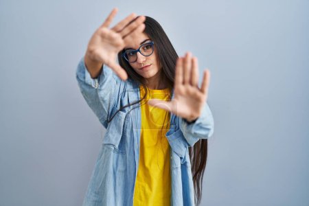 Téléchargez les photos : Jeune femme hispanique debout sur fond bleu faisant cadre en utilisant les mains paumes et les doigts, perspective de la caméra - en image libre de droit