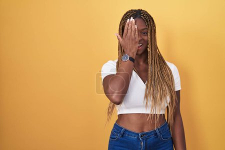 Photo for African american woman with braided hair standing over yellow background covering one eye with hand, confident smile on face and surprise emotion. - Royalty Free Image